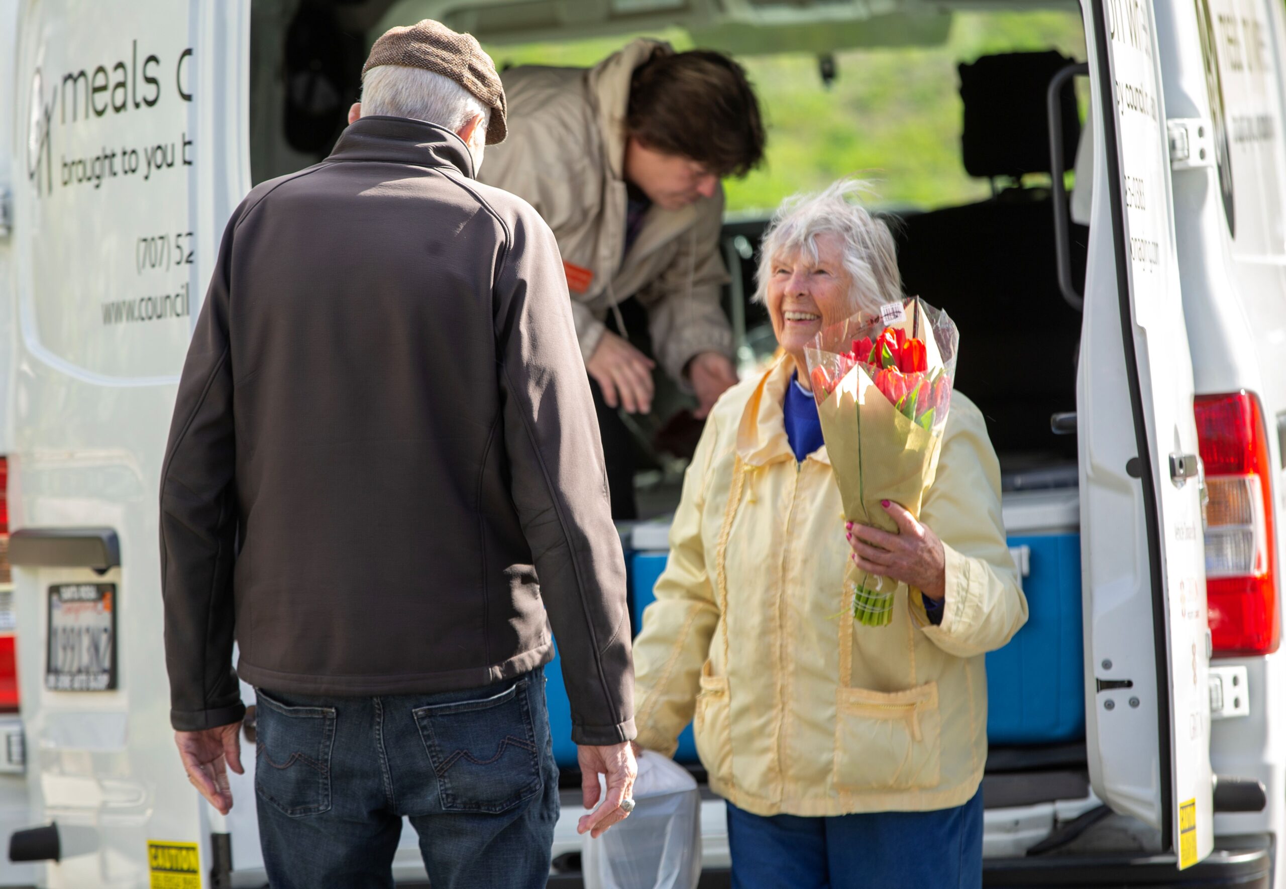 Sabina Blue laughs with Bob Hunter after receiving a bouquet of tulips and a meal from longtime Council on Aging Meals on Wheels driver and Fleet Manager Shannon Holck at the Drive Up/Pick Up site in front of the senior center at King's Valley Senior Apartments in Cloverdale, February 21, 2023. (Chad Surmick / The Press Democrat)